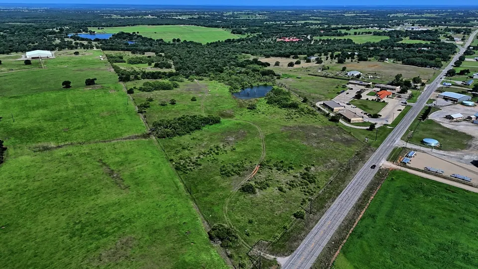 Pastureland with a pond and scattered trees, bisected by a two-lane road near commercial buildings on 67 acres in Parker County